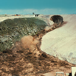 The hole in the dam face continues to enlarge upward near the crest of the dam, the rush of water increases markedly, and erosion cuts deep into bedrock of the abutment.