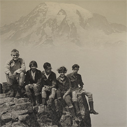 Two women and three men on a mountaineering expedition sit atop Pinnacle Peak at Mount Rainier National Park. The photo, which is part of the Rainier National Park Mountain-Glacier Wonderland Photograph Album, is dated circa 1925.